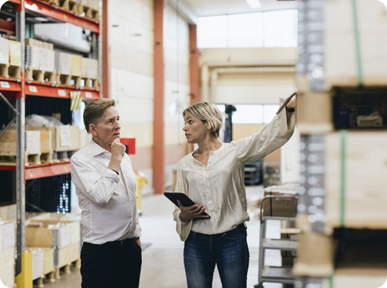 I en ljus lagerlokal pekar en kvinna i vit blus med surfplatta mot hyllor, bredvid står en man i vit skjorta och lyssnar intresserat