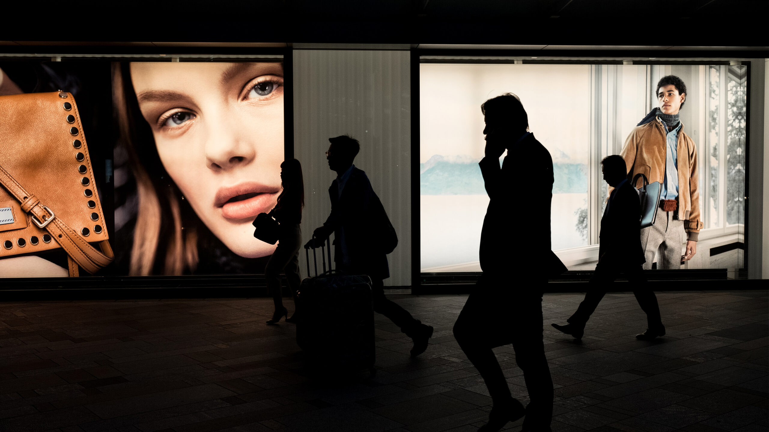 Silhuetter av förbipasserande männsikor framför upplysta reklamskyltar med närbild på kvinna, brun väska och man i moderiktig outfit