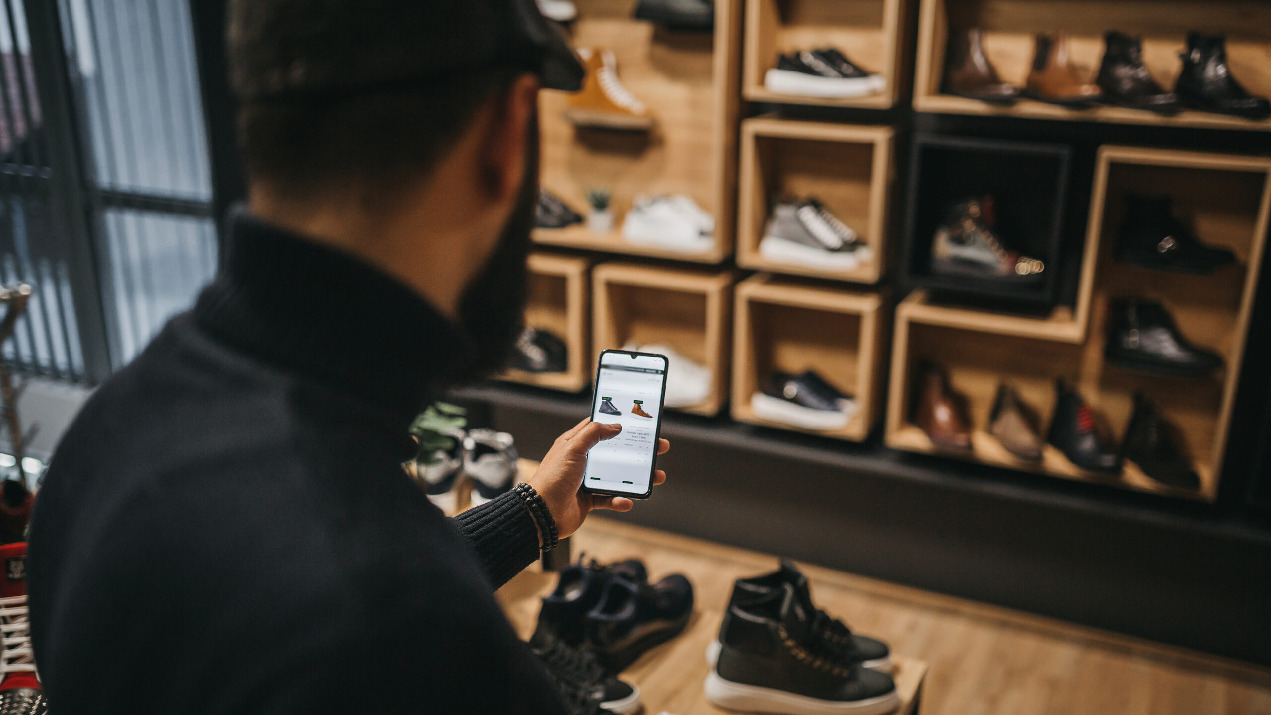 Digital betallösning i mobilen används av person i mörka kläder i en skobutik med träinredning och hyllor fulla av skor