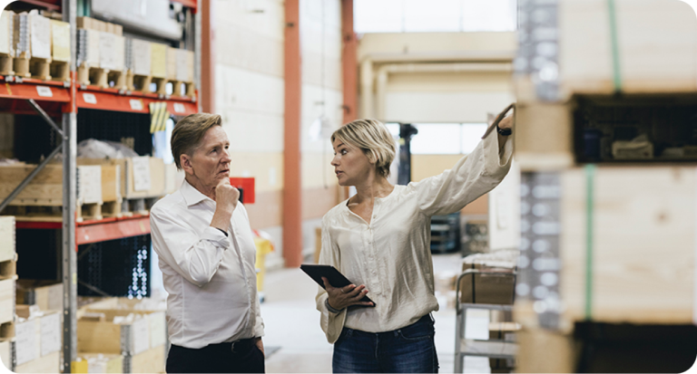 Två personer i ljus lagerlokal; en kvinna i vit blus med surfplatta i handen pekar mot hyllor med varor, medan en man i vit skjorta lyssnar uppmärksamt