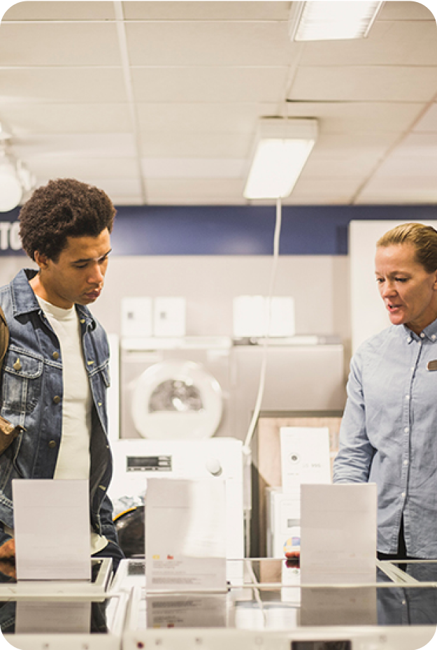 Två personer i en välbelyst modern butik, en manlig kund i blå jeansjacka granskar vitvaror medan en kvinnlig säljare i ljusblå skjorta står bredvid