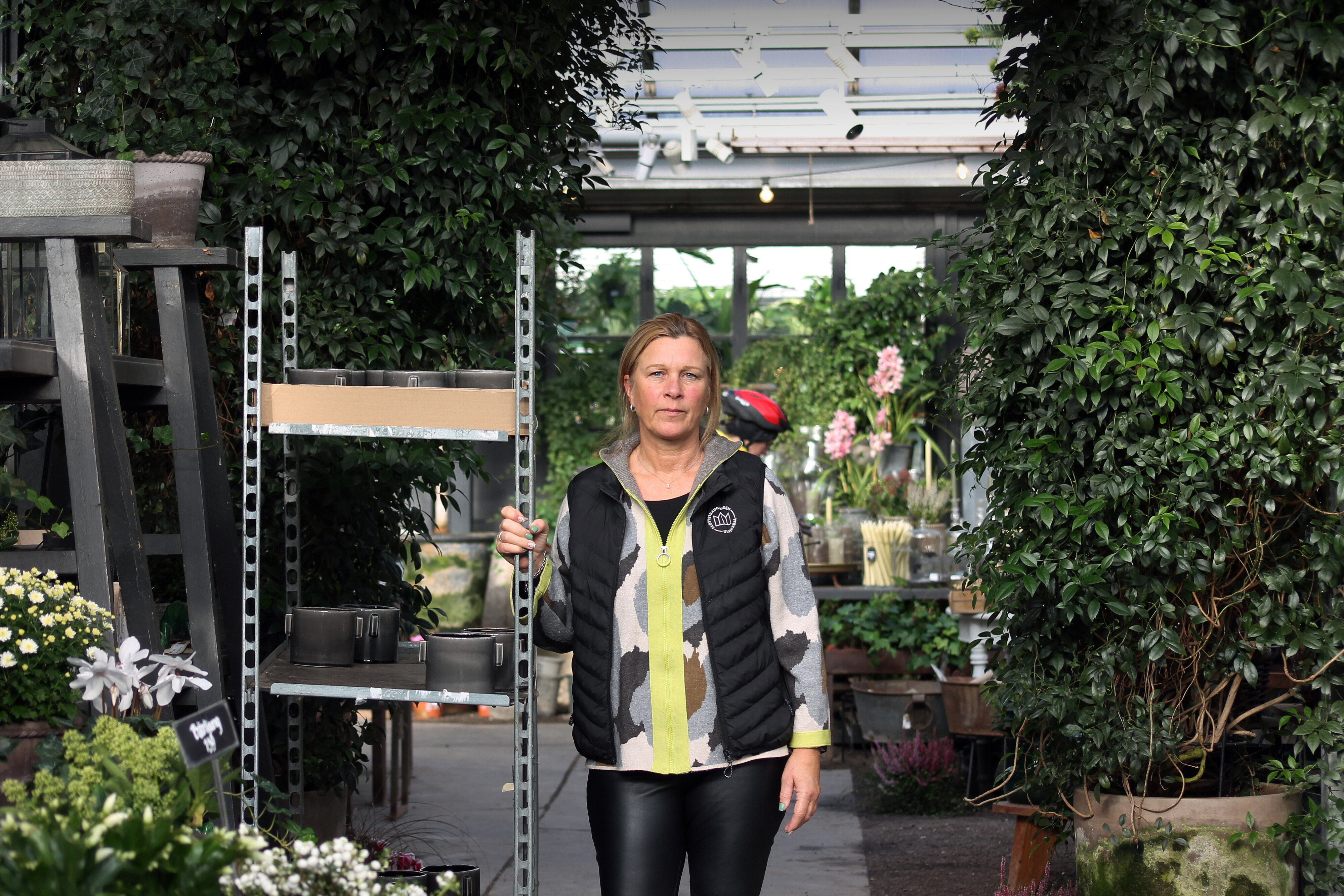 Kvinna i svart väst och mönstrad tröja står i ljust växthus, omgiven av gröna växter, blommor och trädgårdsdetaljer