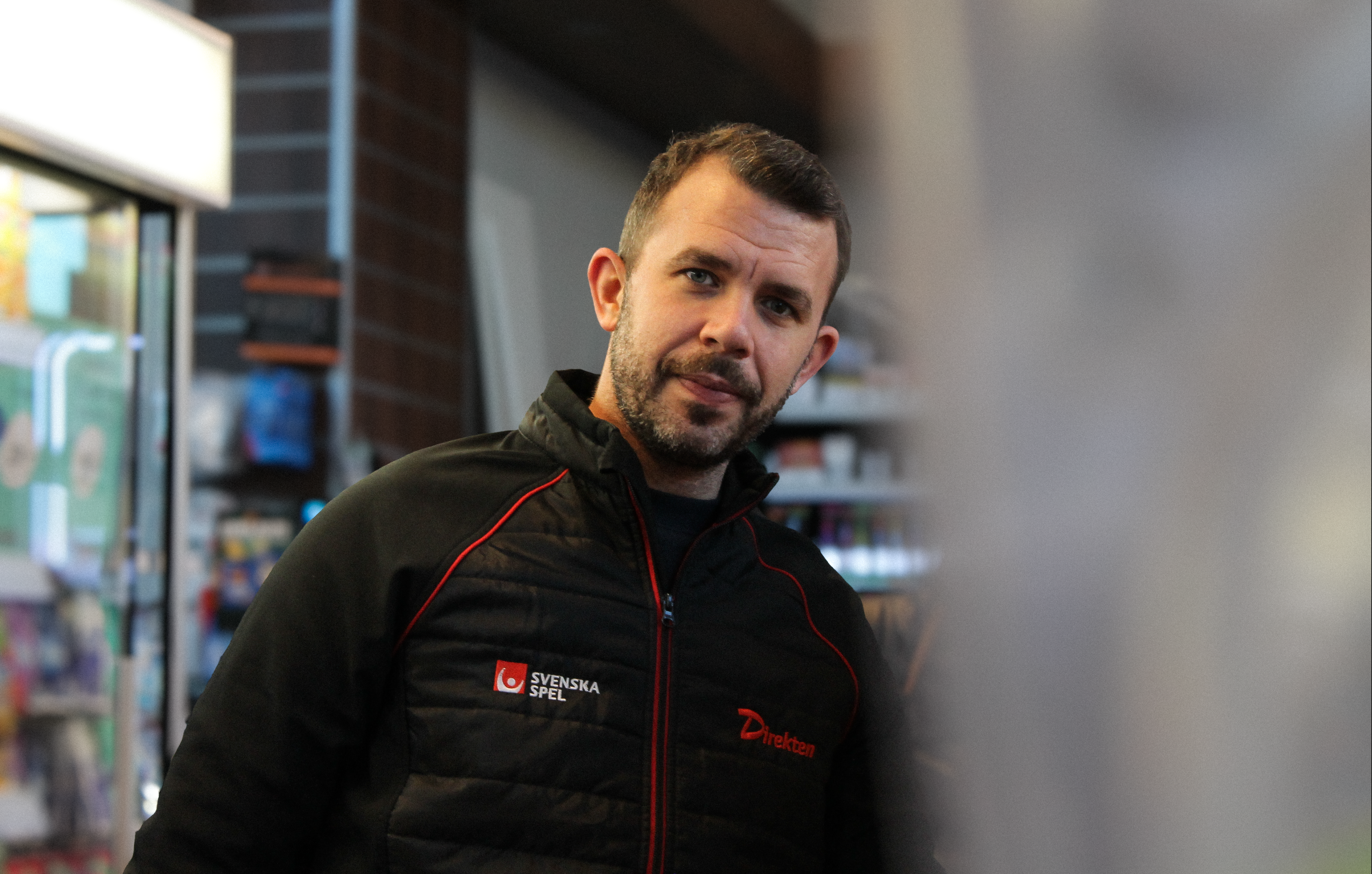 Halvkroppsbild av en man med kort mörkt hår och skägg i svart jacka med röd Svenska Spel-logotyp, stående i butik med suddig bakgrund