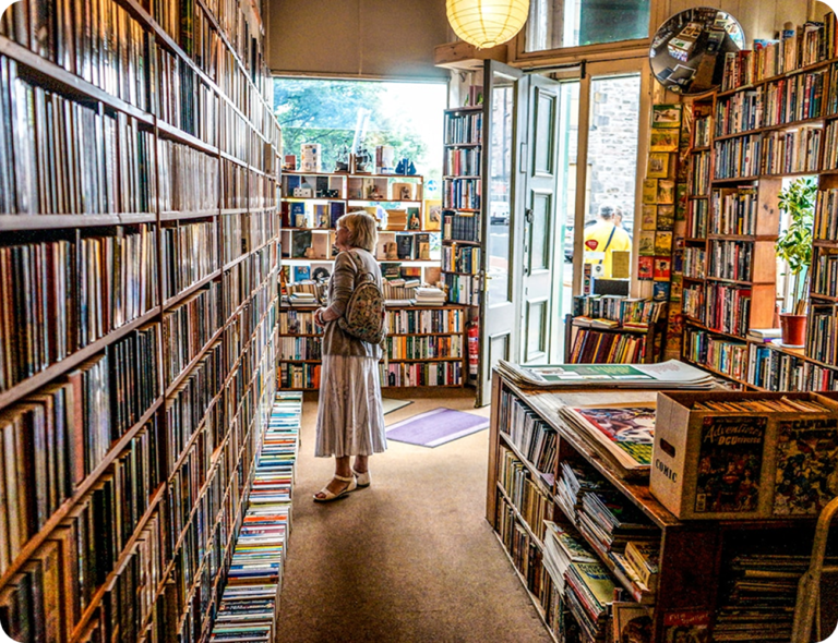 Bokhyllor med färgglada böcker från golv till tak i en charmig, solbelyst bokhandel, där en kund i ljus klänning letar bland böcker