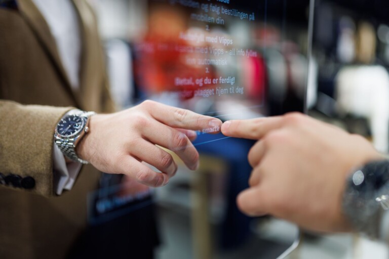 Närbild av Woops digitala pekskärm där en mans hand pekar och speglas i glasytan. Mannen bär en brun ullkavaj i beige och har en klocka på armen