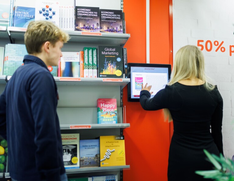 En ung kvinna i svart klänning interagerar med en väggmonterad digital skärm, bredvid till vänster finns en bokhylla med böcker och en man i mörkblå tröja observerar