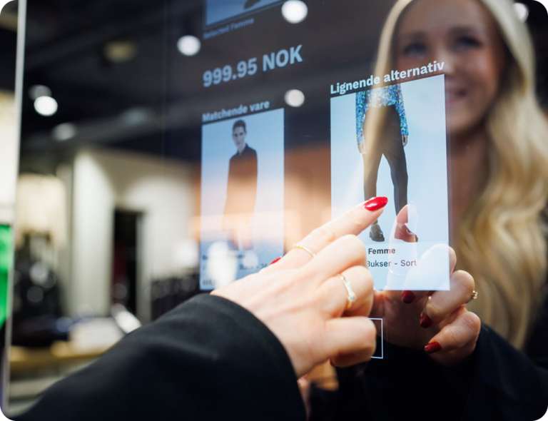 Interaktiv butiksskärm med priser och produktval, en hand med röda naglar pekar på en bild av ett par byxor