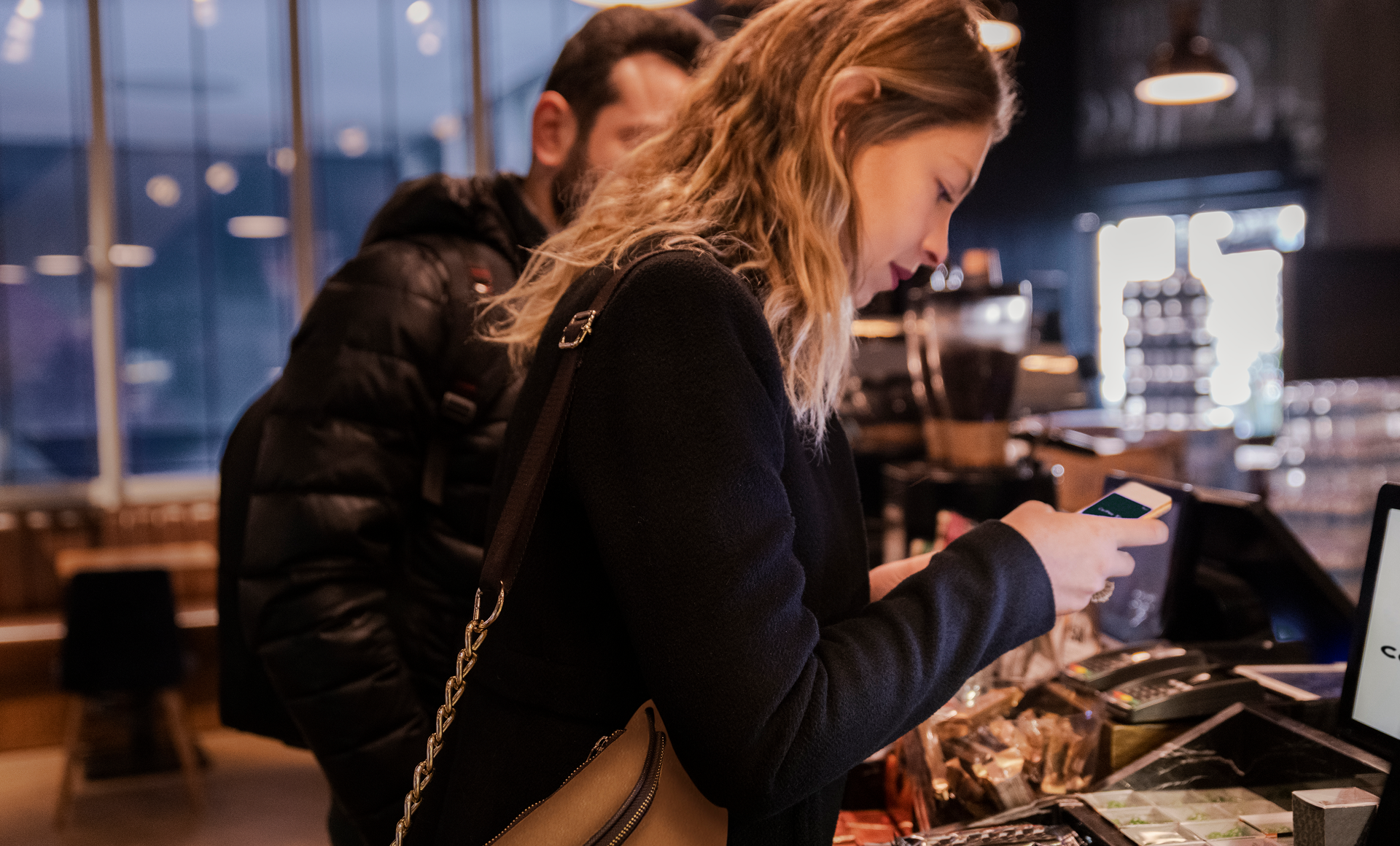 Ung kvinna håller fram mobil för digital betalning i kassan på café eller butik, med en väntande kund bakom i dunkelt belyst butik med mörk inredning