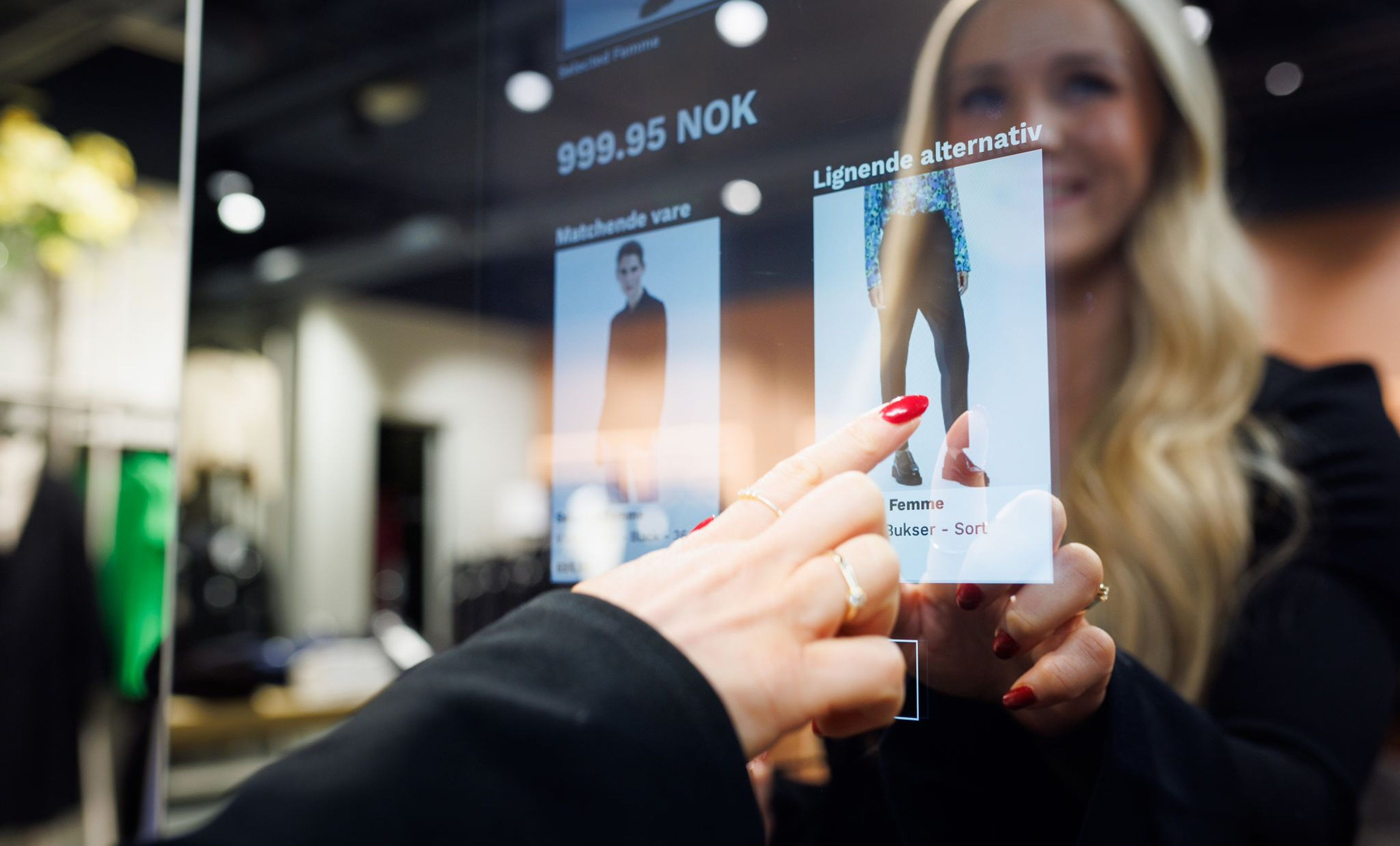 Ung blond kvinna framför en modern interaktiv skärm, där en hand med röda naglar pekar på en produktbild med en bild på ett par byxor