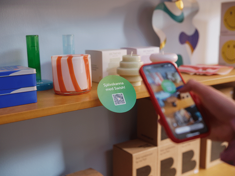 En hand håller i en smart-phone som skannar QR-kod för mobilbetalning med Swish på butikshylla med färgglada vaser och ljuslyktor